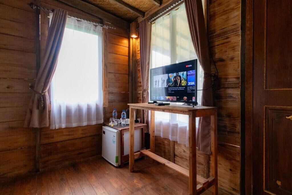 Habitación de cabaña de madera con una televisión sobre una mesa de madera, un refrigerador pequeño y una ventana con cortinas.