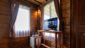 Habitació d'un xalet de fusta amb televisió sobre una taula de fusta, un frigorífic petit i una finestra amb cortines.