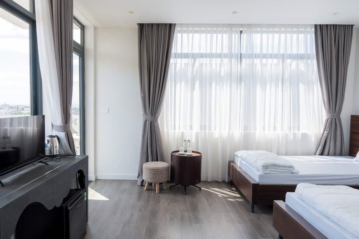 Una habitación de hotel con cama, una mesita redonda con lámpara, televisión y ventana con cortinas.