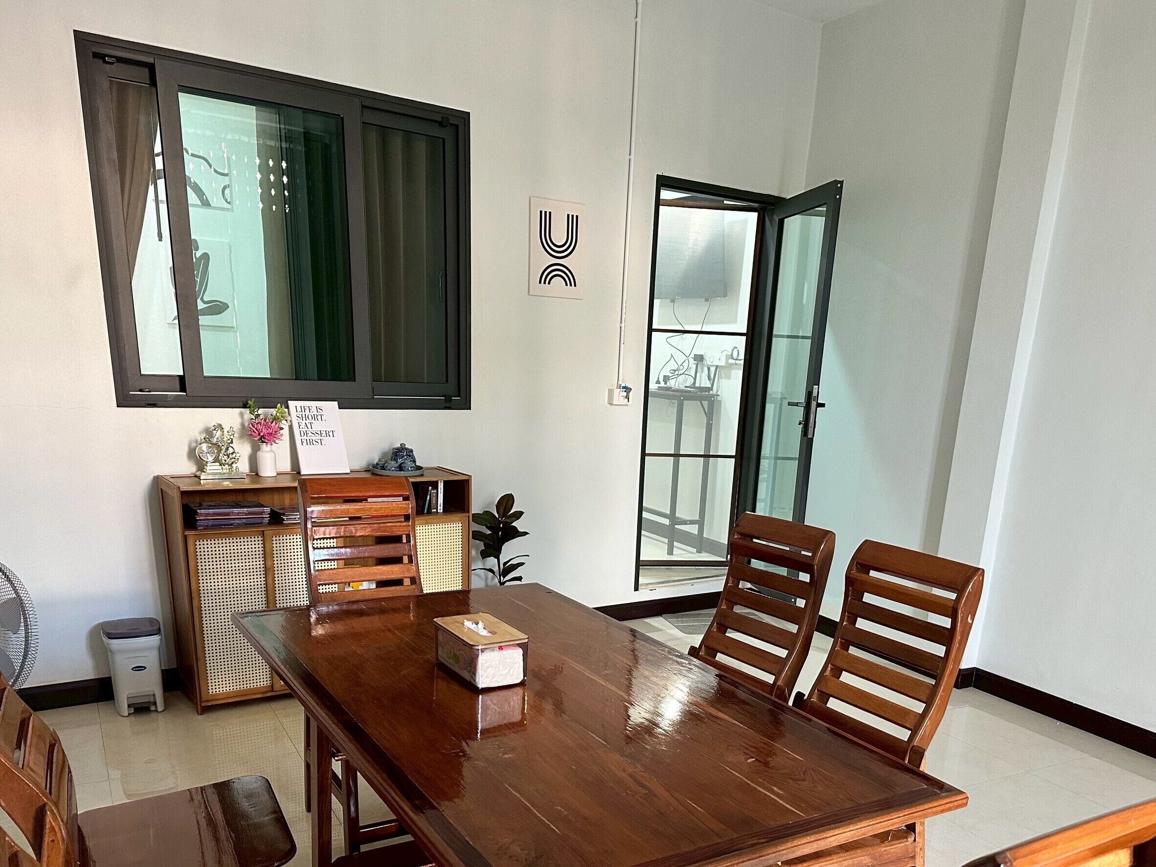 Een eetgedeelte met een houten tafel, vier stoelen, een bijzettafeltje met decoratieve objecten en een schuifdeur naar een balkon.