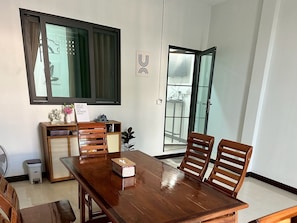 A dining area with a wooden table, four chairs, a small side table with decorative items, and a sliding glass door leading to a balcony. - Cocoon Casa (Hat Yai)