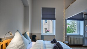 Een hotelkamer met een bed, bedlampen, een raam met uitzicht op gebouwen, een radiator en een klein tafeltje.