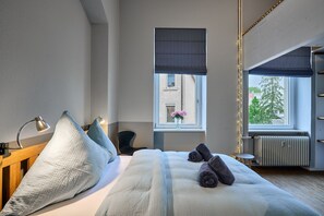 A hotel room with a bed, bedside lamps, a window with a view of buildings, a radiator, and a small table. - Loft | Altstadt Urban | seeglueck (Konstanz)