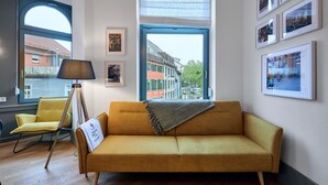 A room with a sofa, a chair, a floor lamp, and a large window with a view of buildings. - Studio | Old Town | seeglueck (Konstanz)
