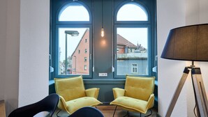 A room with two yellow chairs, a black chair, a floor lamp, and a large window with a view of buildings outside. - Studio | Old Town | seeglueck (Konstanz)