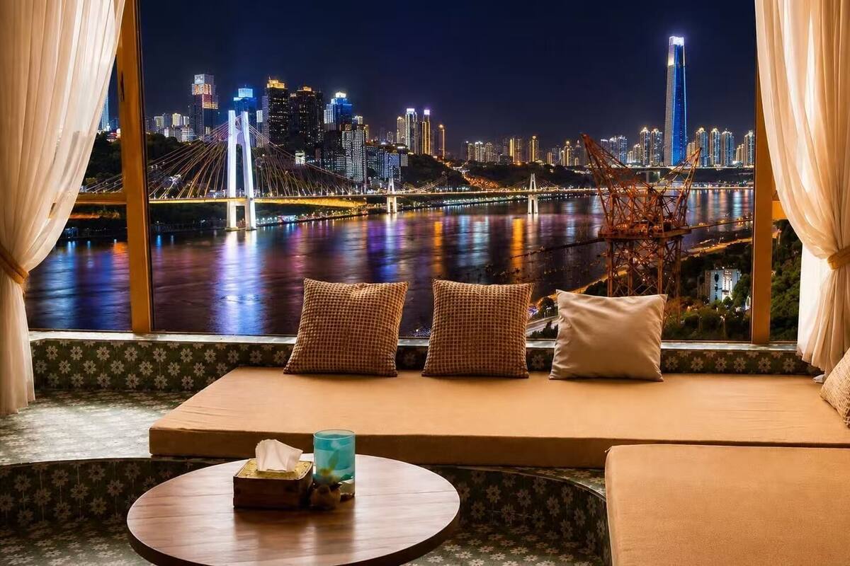 Ein Zimmer mit Blick auf eine Stadtlandschaft, ein Sofa mit Kissen, ein kleiner Tisch mit Taschentuchbox und Glas sowie Vorhänge.