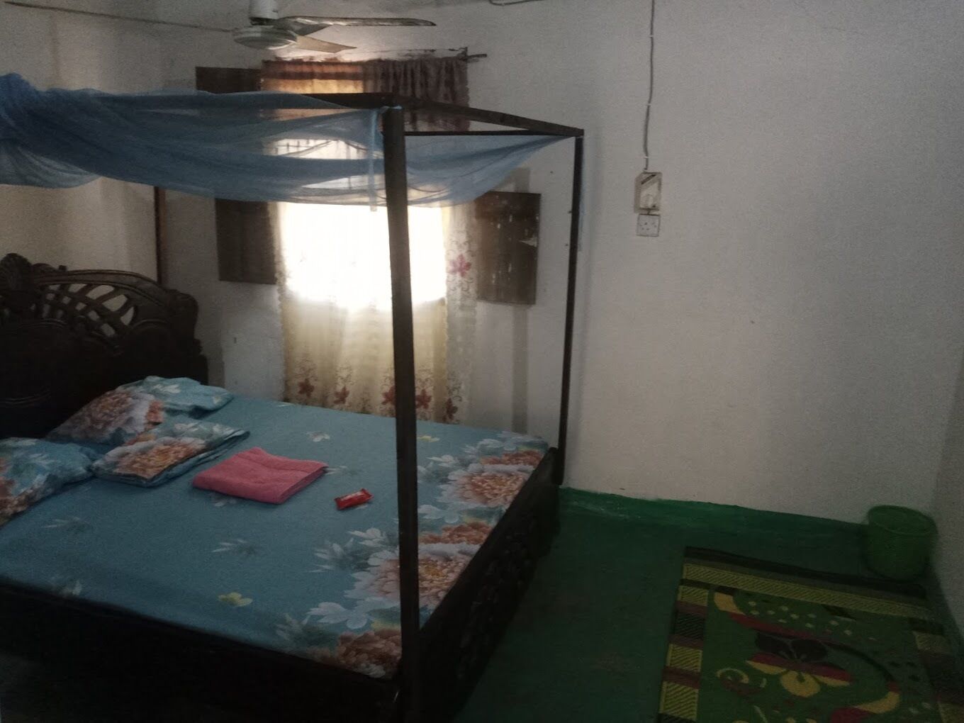 A single bed with a mosquito net, a floral bedspread, and a green mat beside it.