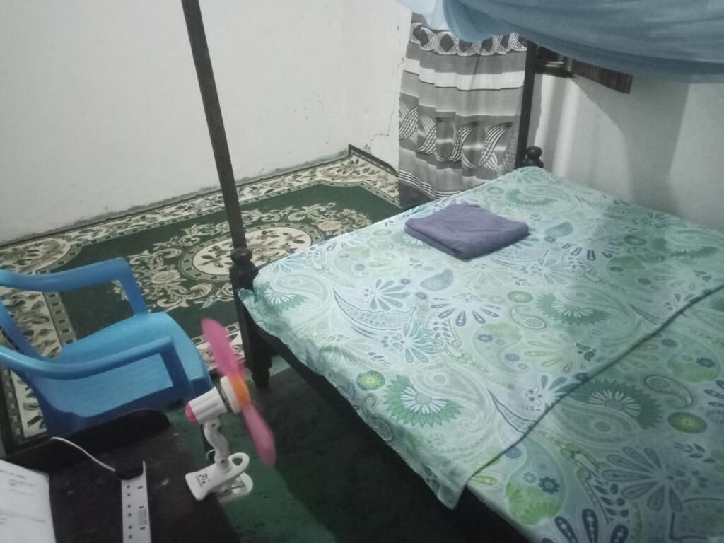 Ein kleines Zimmer mit einem Bett, einem blauen Stuhl, einem Ventilator und einem gemusterten Teppich.