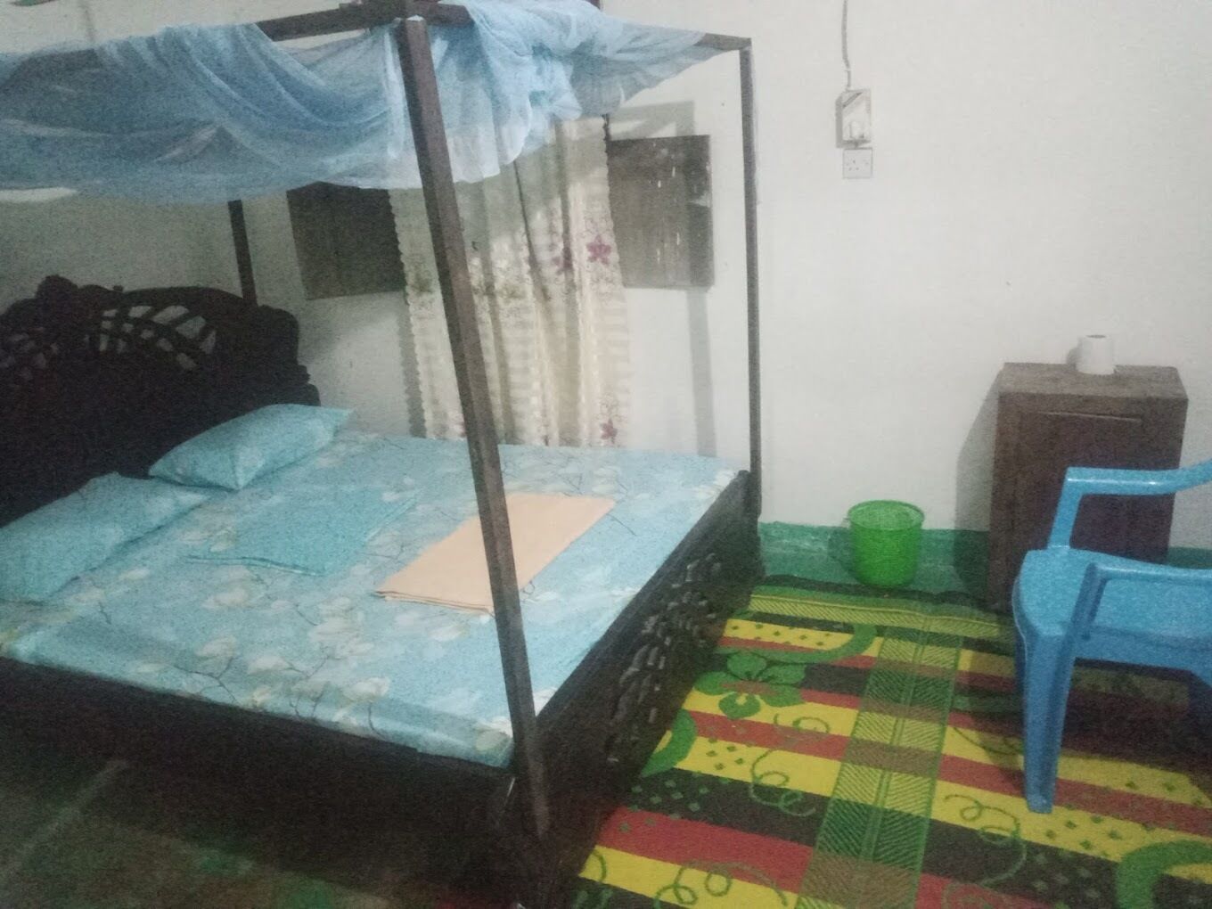 A single bed with a canopy, a small desk, a chair, and a green bucket.