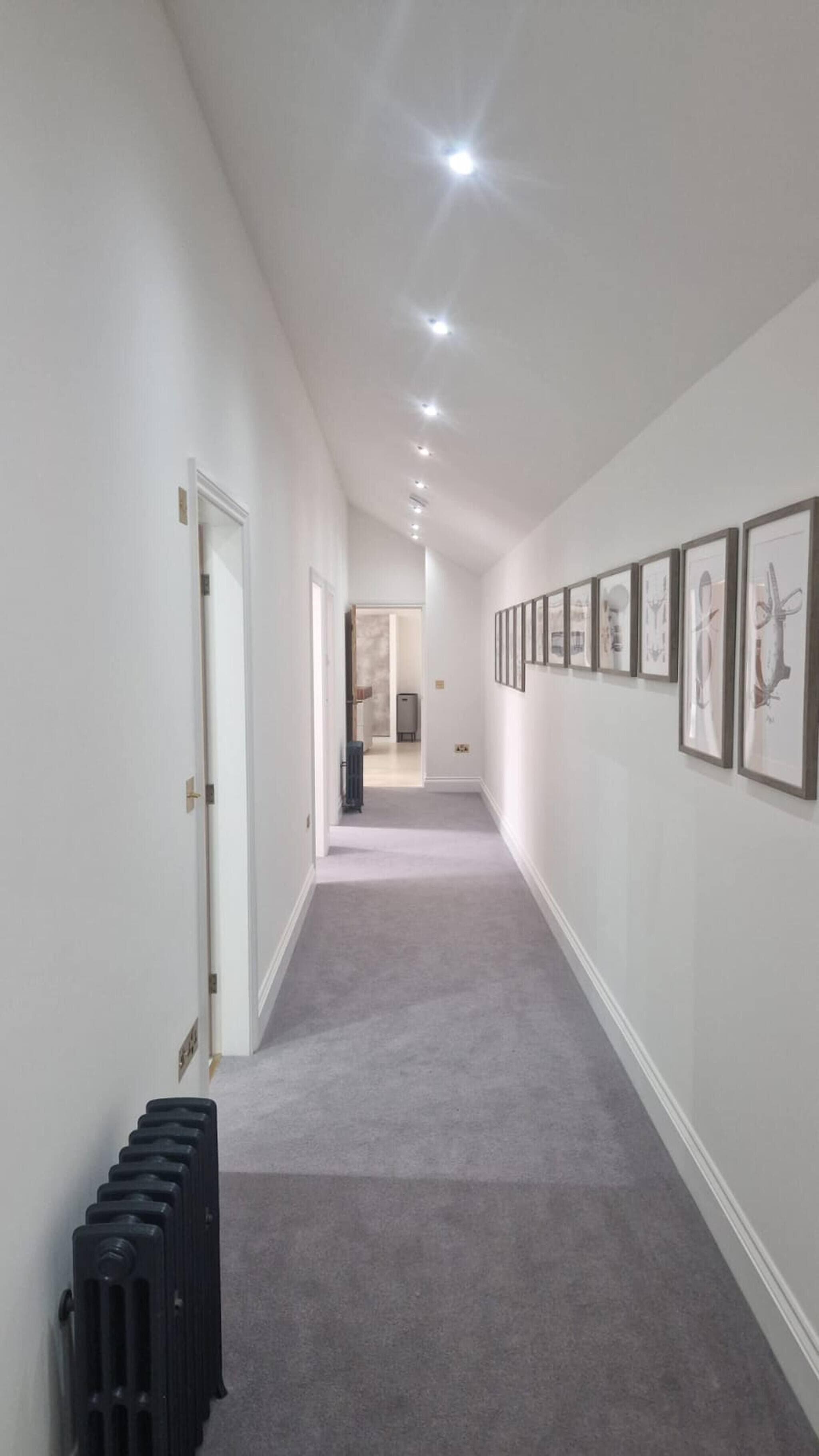 A hallway with a radiator, framed artwork on the walls, and recessed lighting.