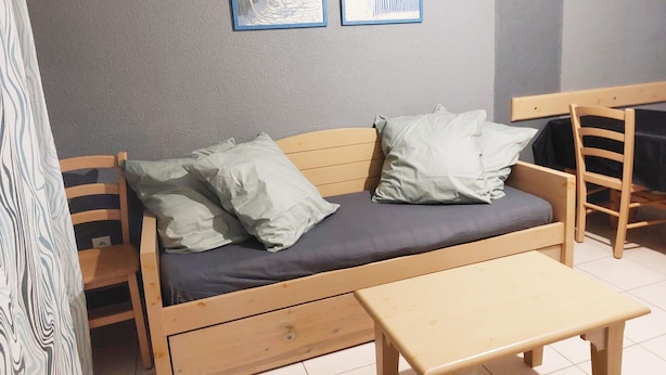 Ein Zimmer mit einem hölzernen Bett, zwei Kissen, einem kleinen Tisch und zwei gerahmten Bildern an der Wand.