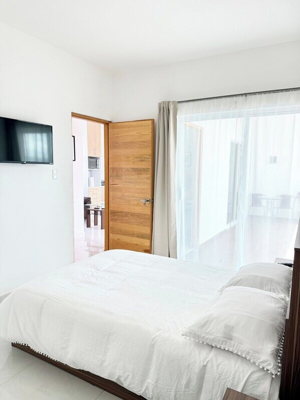A bedroom with a bed, a wooden door, a TV mounted on the wall, and a window with curtains. - Charming 2BR Apt in Oaxaca (Oaxaca de JuÃ¡rez)