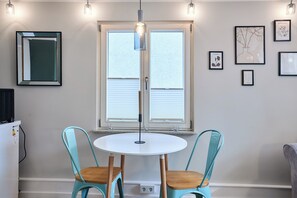 Une pièce avec une table ronde, deux chaises, une grande fenêtre avec des stores, un luminaire suspendu et des œuvres d’art encadrées accrochées aux murs.
