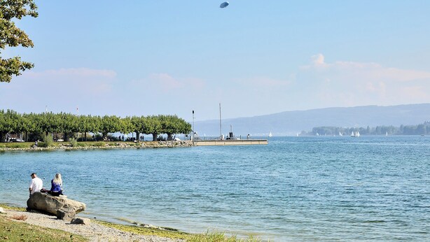 Ein ruhiger See mit einer Anlegestelle, Booten und einem wolkenlosen Himmel.
