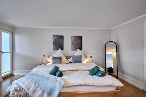Un lit bien fait, avec des oreillers et une couverture, une table de chevet avec une lampe, un grand miroir et deux œuvres d’art accrochées au mur.