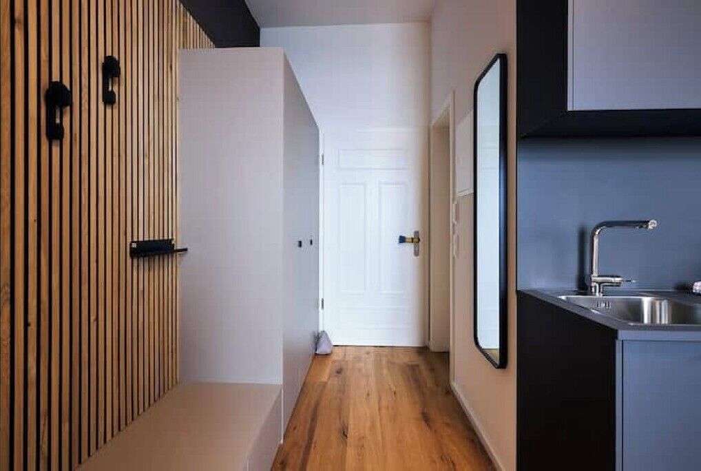 Un couloir étroit, avec un lambris en bois sur un côté, une porte blanche au fond, et un coin cuisine avec un évier et des placards sur la droite.