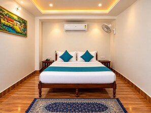 A hotel room with a bed, a nightstand, an air conditioning unit, a wall-mounted fan, and a painting on the wall. - Wild Vista Marchula Riverside Resort Jim Corbett (Marchula)