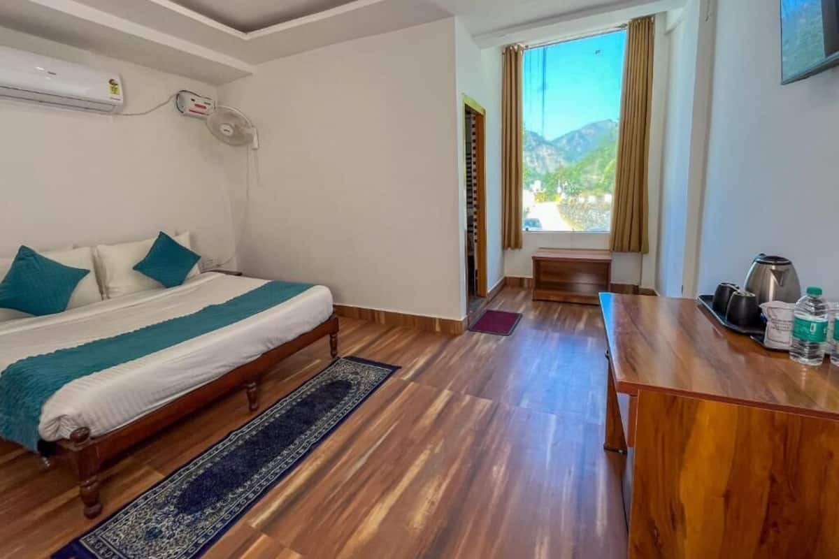 A hotel room with a bed, wooden floor, a desk with a kettle, and a window with curtains.