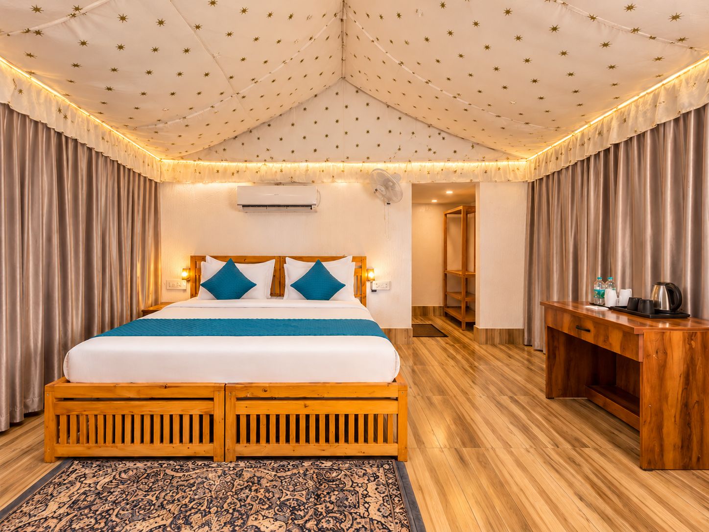 A double bed with a wooden frame, a headboard with two blue pillows, a bedside table with a kettle, and a patterned rug.