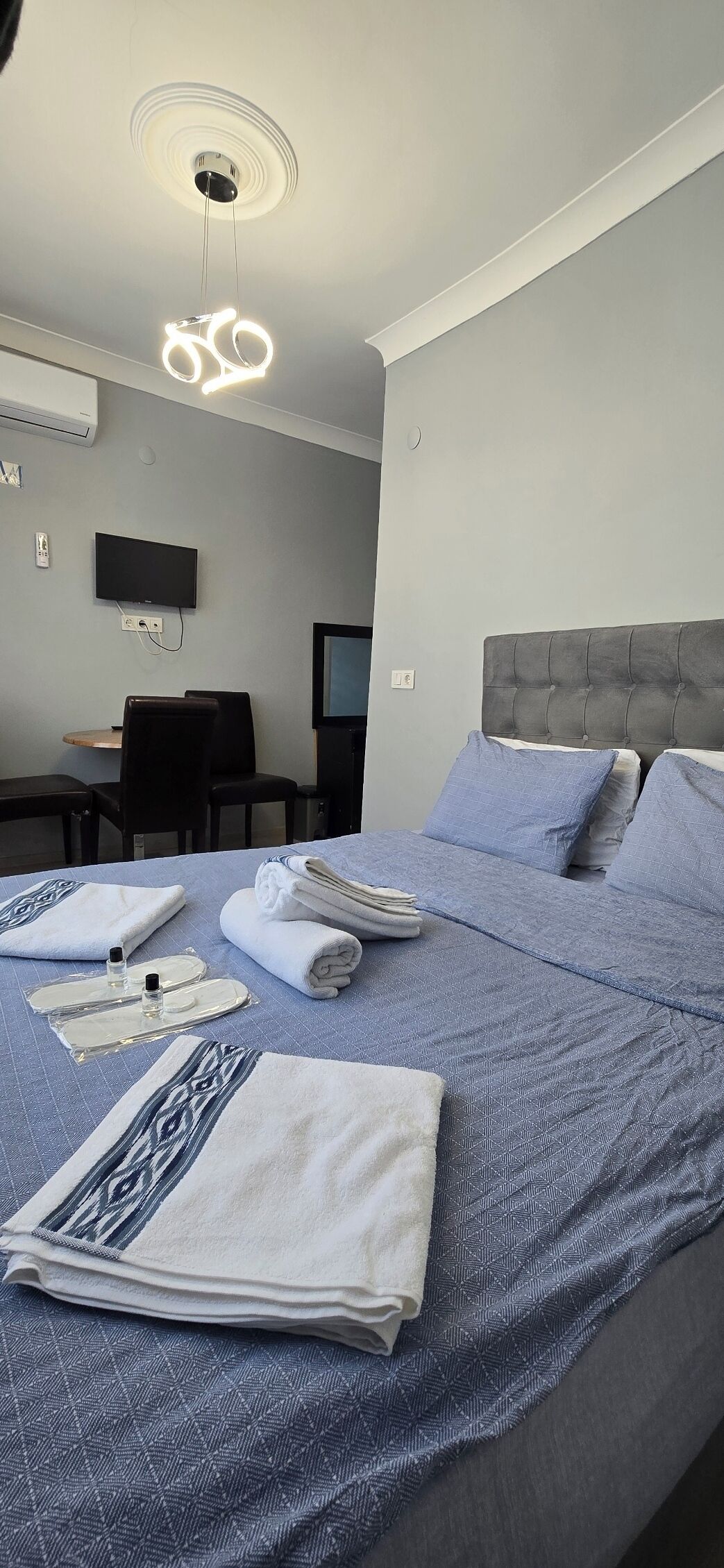Une chambre d’hôtel avec un lit soigneusement fait, un téléviseur à écran plat fixé au mur, un bureau avec une chaise et un luminaire moderne au plafond.