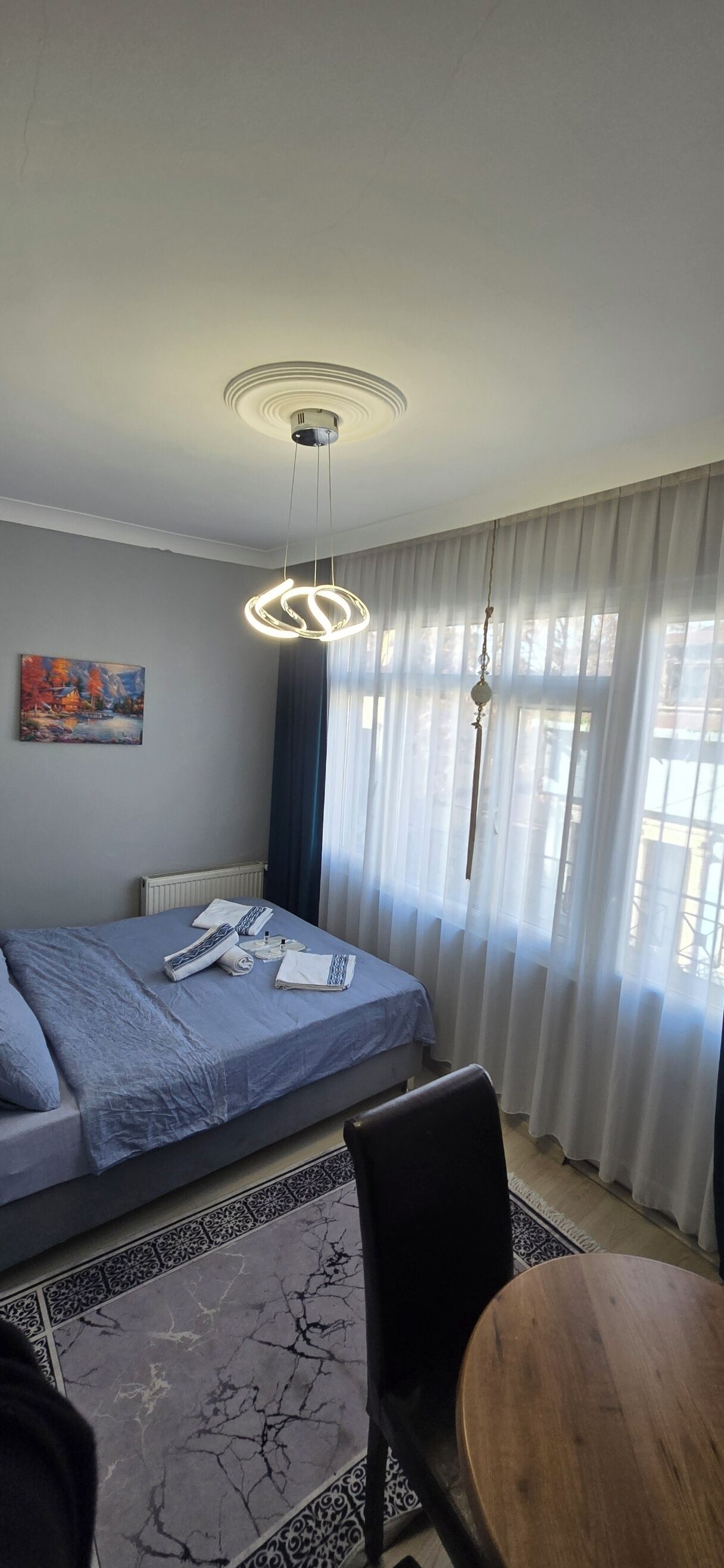 A hotel room with a bed, a table, a chair, a painting on the wall, and a window with curtains.