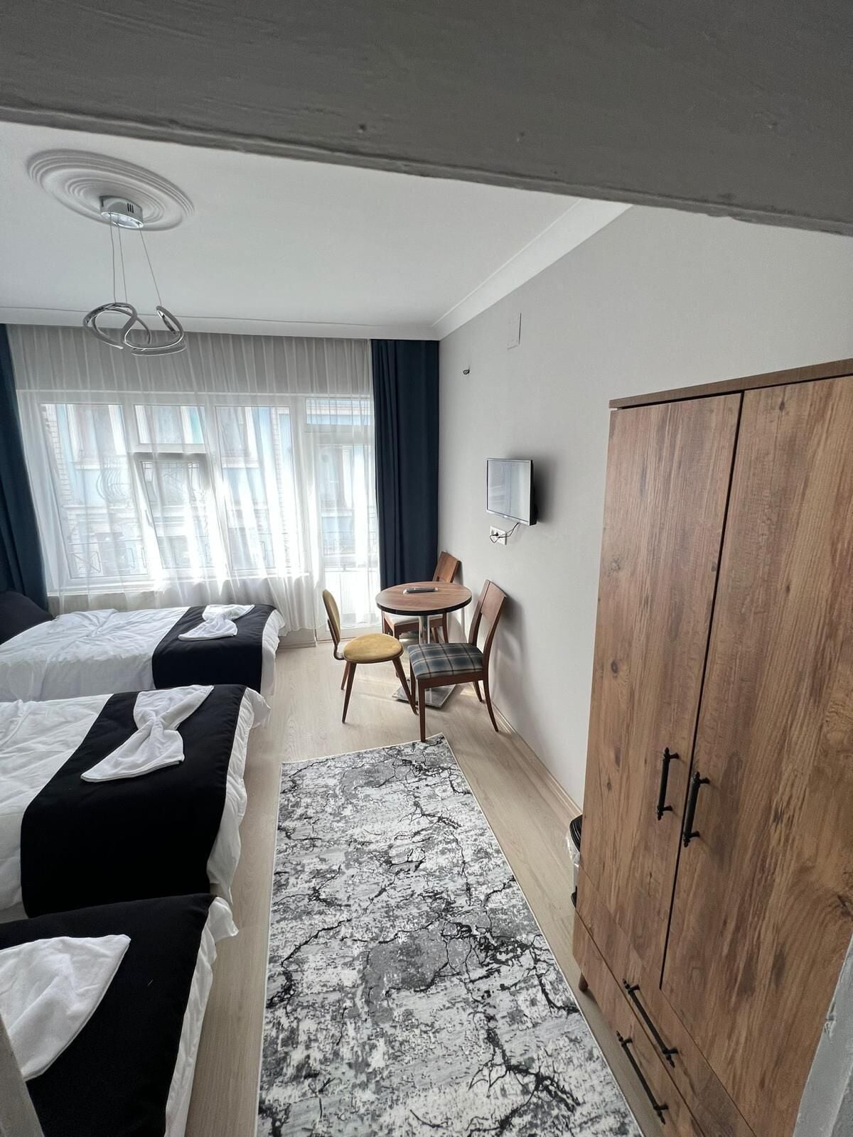 A hotel room with two beds, a dining table, chairs, a wooden wardrobe, and a television on the wall.