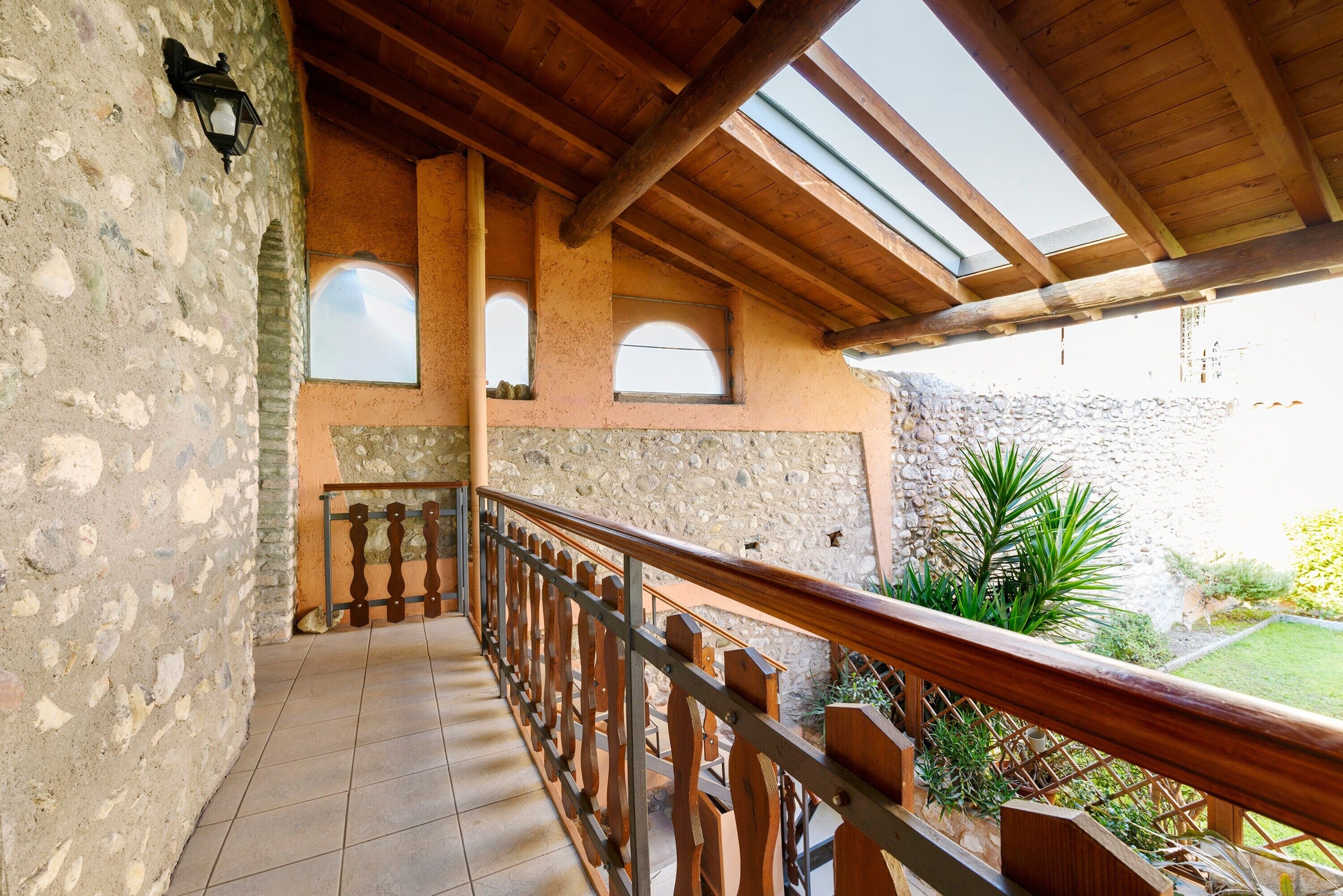 A balcony with a wooden railing, stone walls, and a view of a garden.