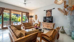 A spacious living room with a sofa set, a coffee table, and a television. There is a large window providing a view of greenery outside. - Private Pool | Daily Housekeeping | Central Seminyak (Seminyak)