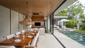 Un comedor moderno con una larga mesa de madera dispuesta para seis personas, sillas blancas acolchadas y una gran lámpara colgante tejida. Un televisor de pantalla plana está montado en la pared sobre la mesa.