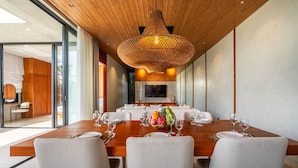 A modern dining area with a wooden table set for six, surrounded by upholstered chairs. A large woven light fixture hangs from the ceiling, and a flat-screen TV is mounted on the wall behind the table. - Modern Villa | Private Pool | Near Canggu Beach (Kerobokan)