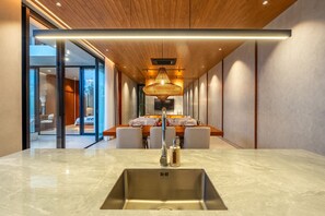 A modern kitchen with a large island, a built-in sink, and a dining area with a wooden table and chairs. - Modern 3BR Pool Villa | Green views | Near Berawa (Kerobokan)