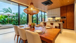 Un espace de repas moderne avec une table en bois dressée pour six personnes, un luminaire suspendu et une vue sur une végétation luxuriante à l’extérieur.
