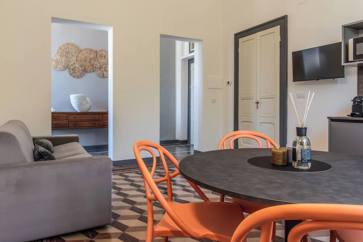 A modern living room with a grey sofa, orange chairs, a round table, and a TV mounted on the wall.