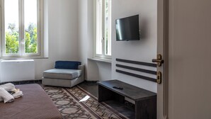 A hotel room with a bed, a small seating area, a TV mounted on the wall, and a window providing a view of greenery outside. - Villa Leda (Porto Recanati)