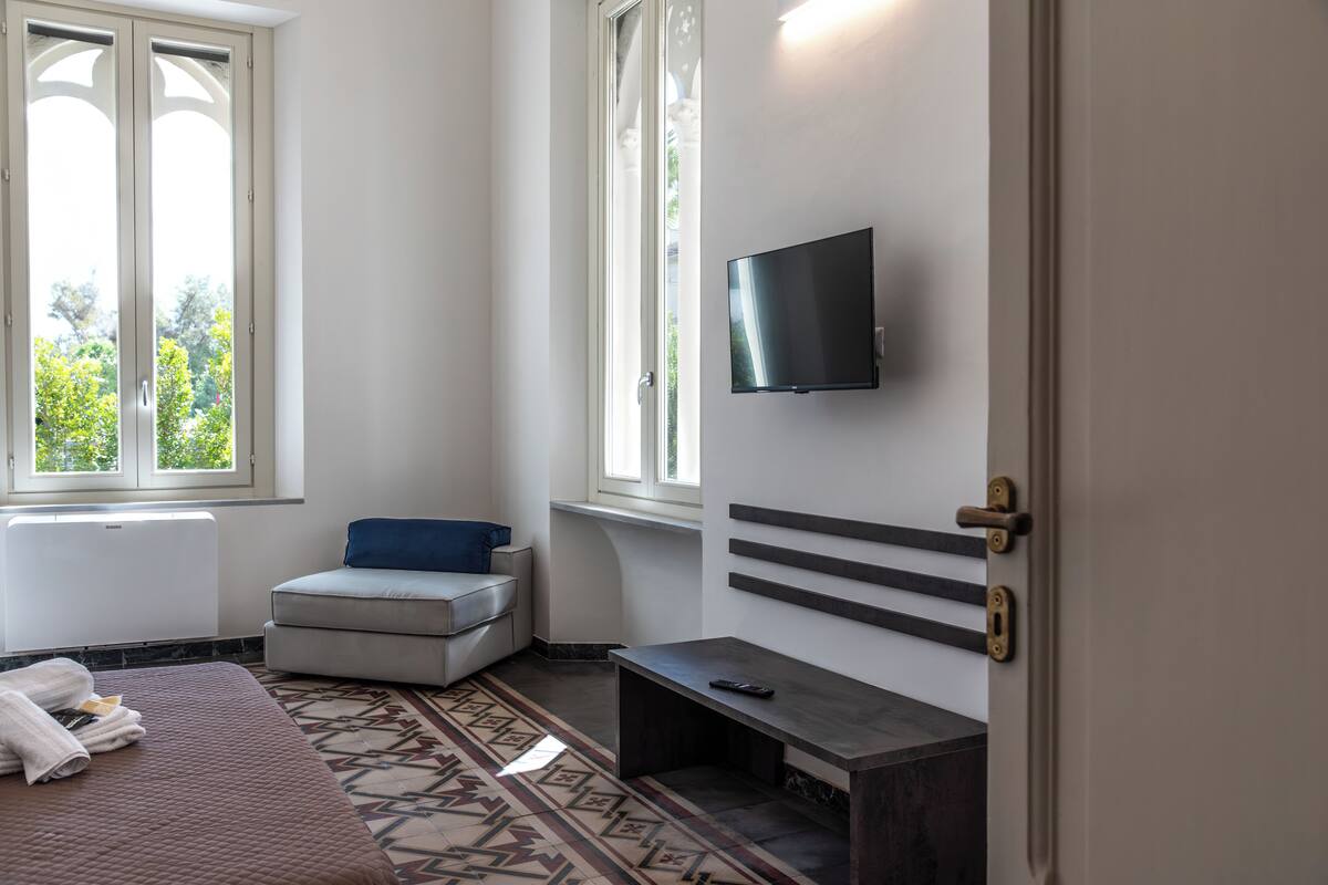 A hotel room with a bed, a small seating area, a TV mounted on the wall, and a window providing a view of greenery outside.