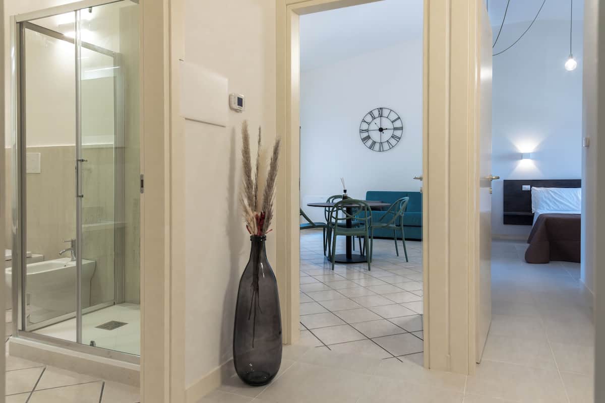 A modern hotel room with a glass shower enclosure, a vase with pampas grass, a dining area with a clock on the wall, and a bed with a white headboard.