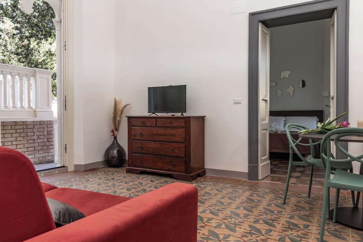 A room with a wooden dresser, a television on top, a red sofa, a dining table with chairs, and a patterned floor.