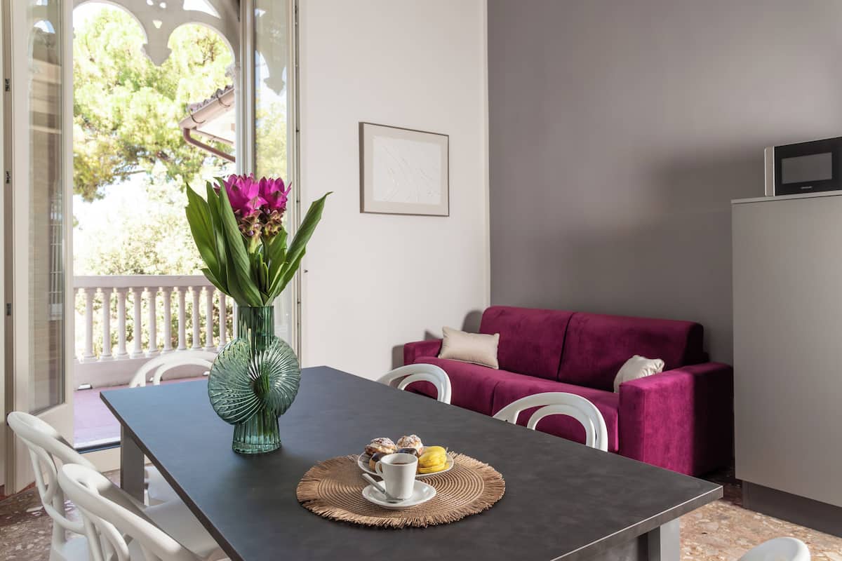 A dining area with a table set for two, a vase with flowers, and a view of a balcony with a railing.