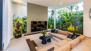 Een moderne woonkamer met een beige bank, een zwarte salontafel en een grote televisie. Achter de bank bevindt zich een glazen wand met planten.
