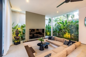 Ein modernes Wohnzimmer mit einem beigefarbenen Sofa, einem schwarzen Couchtisch und einem großen Fernseher. Eine Glaswand mit Pflanzen befindet sich hinter dem Sofa.