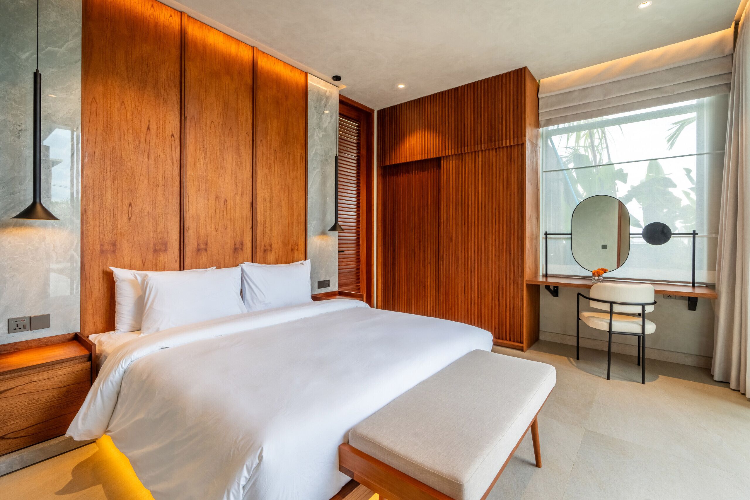A modern bedroom with a large bed, wooden paneling, a bedside table, a chair, a window with a view, and a mirror.