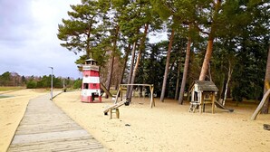 Eine Holzterrasse führt zu einem Sandstrand mit einem rot-weißen Leuchtturm, einer Schaukel und einem kleinen Holzspielhaus.