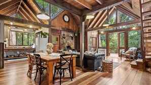 Interior rústico de una cabaña con comedor, chimenea y ventanas amplias que dan al bosque.