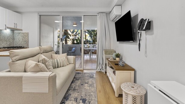A modern living room with a sofa, a coffee table, and a TV mounted on the wall. There is a sliding glass door leading to a balcony with outdoor furniture. - Bright 1-Bed Apartment in Fortitude Valley (Fortitude Valley)