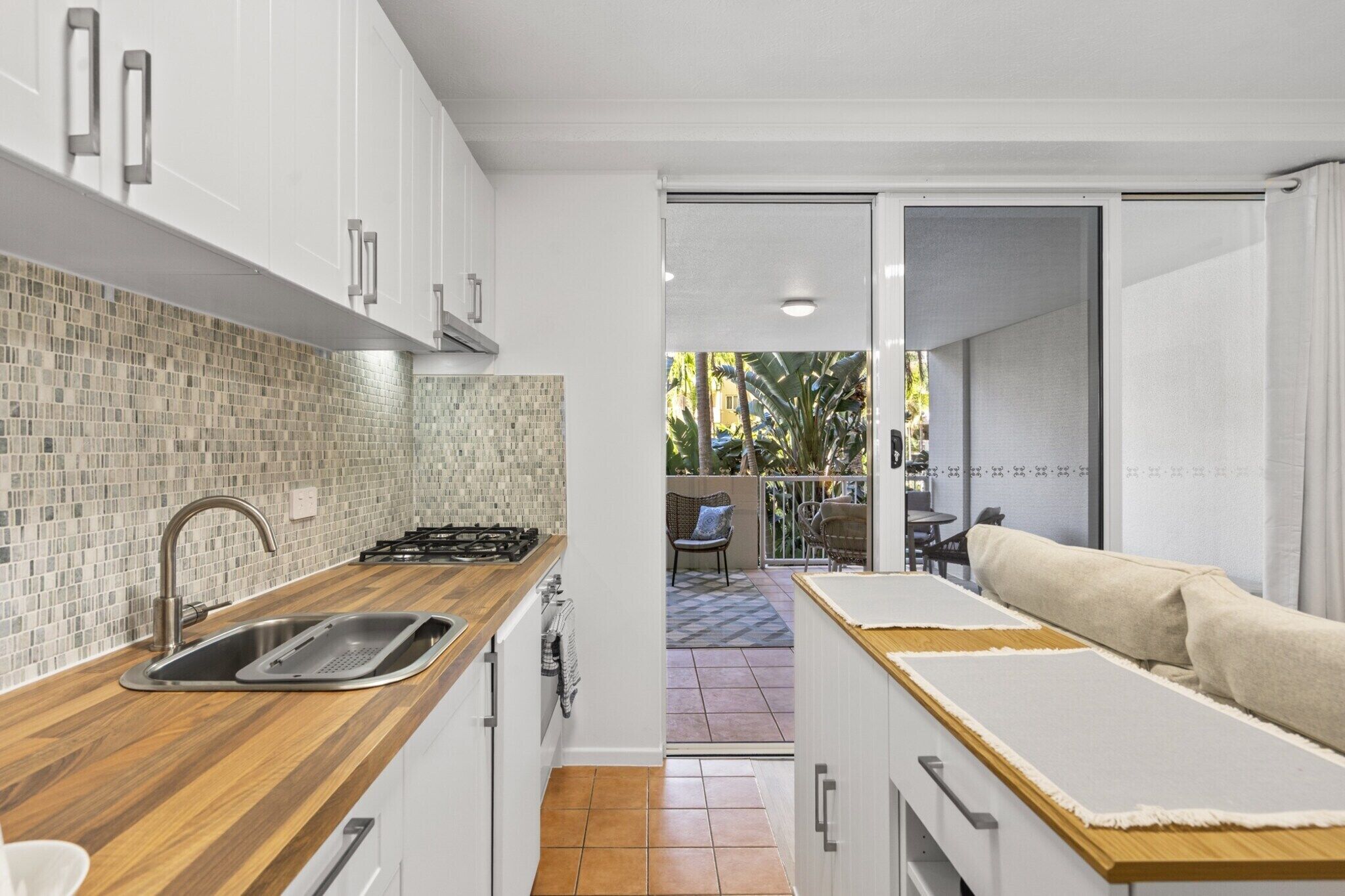 Una cocina moderna con encimera de madera, pileta de acero inoxidable y alacenas blancas. Hay una puerta corredera que da a un espacio exterior con una silla y una mesa.