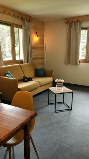 Une pièce chaleureuse avec un canapé, une petite table et une chaise. Il y a des fenêtres avec des rideaux, un luminaire fixé au mur et un appareil photo posé sur le canapé.
