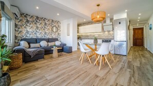 A modern living room with a sofa, coffee table, and dining area. There is a kitchen with a refrigerator, stove, and sink. The room has wooden flooring and a tropical wallpaper design. - Venecia Beach Apartment Javea Arenal (Javea / Xàbia)