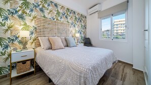 A bedroom with a bed, nightstands, a lamp, a chair, a window, and a tropical wallpaper. - Venecia Beach Apartment Javea Arenal (Javea / Xàbia)