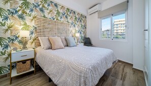 Ein Schlafzimmer mit einem Bett, Nachttischen, einer Lampe, einem Sessel, einem Fenster und tropischem Wandpapier.