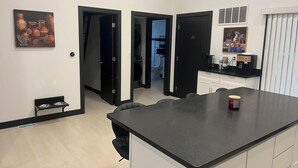Een moderne keuken met een centraal eiland, zwarte aanrechtbladen en een koffieapparaat. Er zijn twee deuren die naar andere ruimtes leiden, en een klein plankje met kommen.